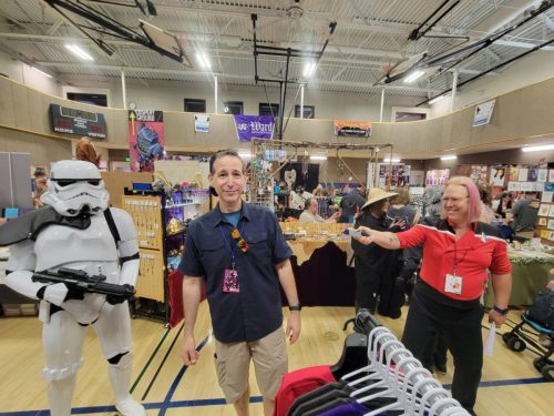 Sarah in a red Lower Decks uniform and a Stormtrooper capture an unsuspecting convention goer at phaser/rifle-point.